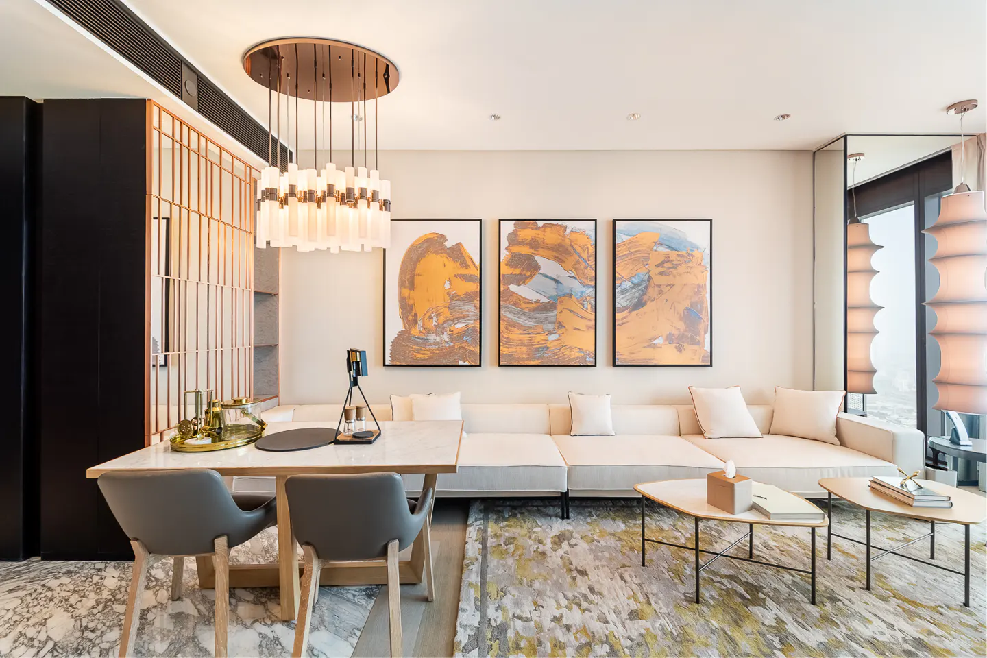 Bright living room with a white sofa, abstract art, and a dining table with gray chairs. A modern chandelier hangs above the table.
