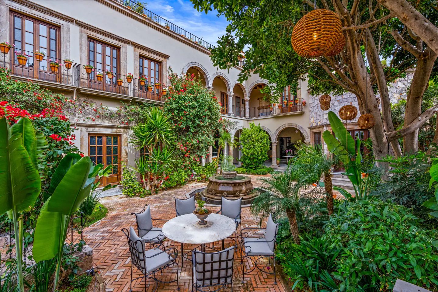 Courtyard view of a two-story building with a fountain, table, and chairs. The building has arched walkways and flower boxes on the balconies.