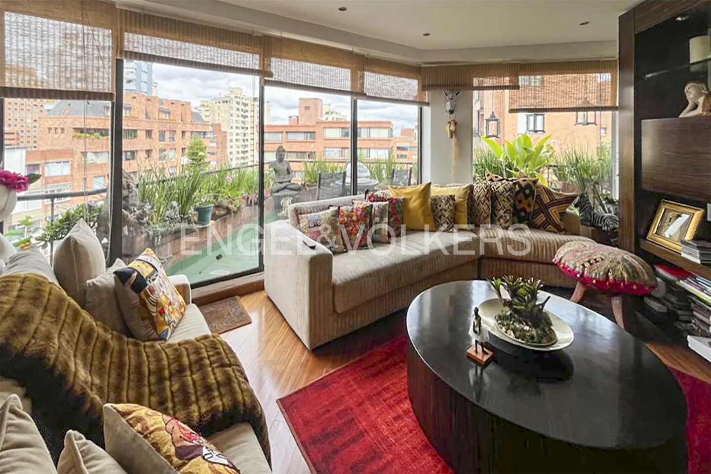 A bright living room with sofas, a dark wood coffee table, and a red rug. A balcony with plants and a Buddha statue is visible through the window.