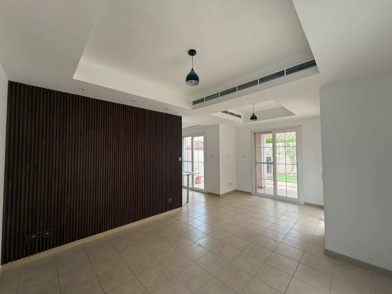 An empty living room with beige tile floors, white walls, and a dark wood accent wall. Two pendant lights hang from the ceiling. Glass doors lead to a green yard.