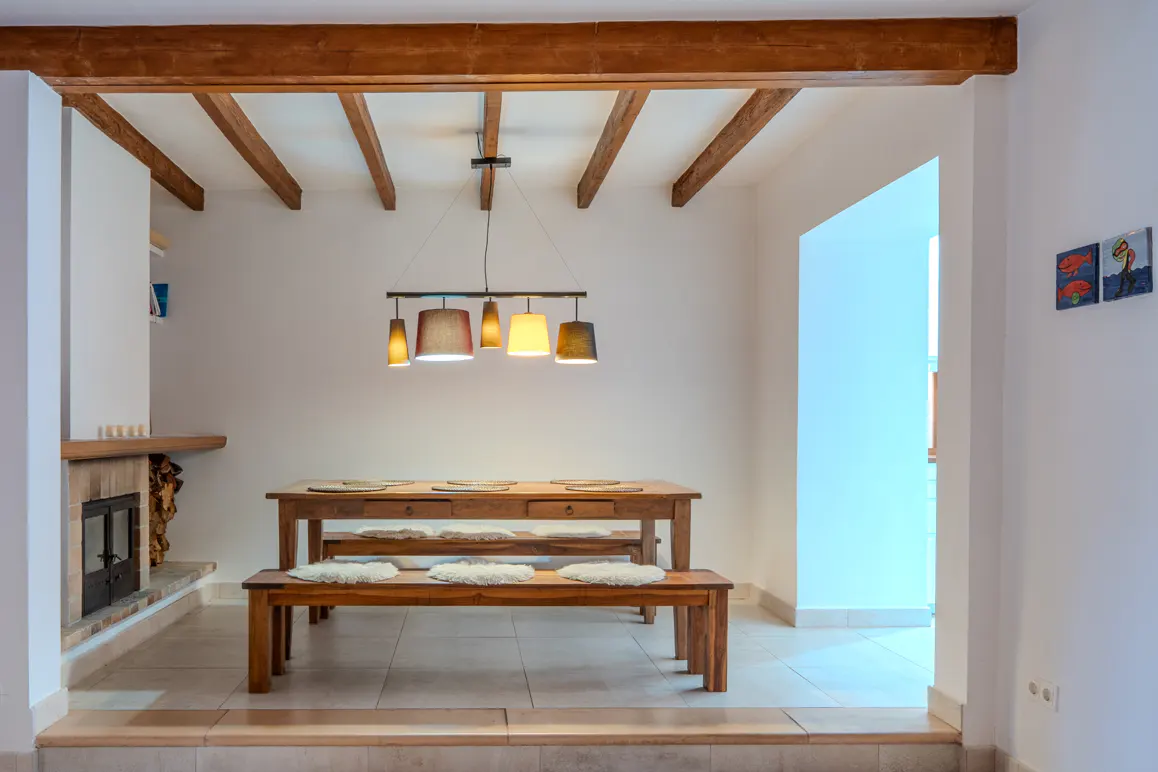 Dining area with wooden table and benches, white walls, exposed ceiling beams, and a multi-pendant light fixture.