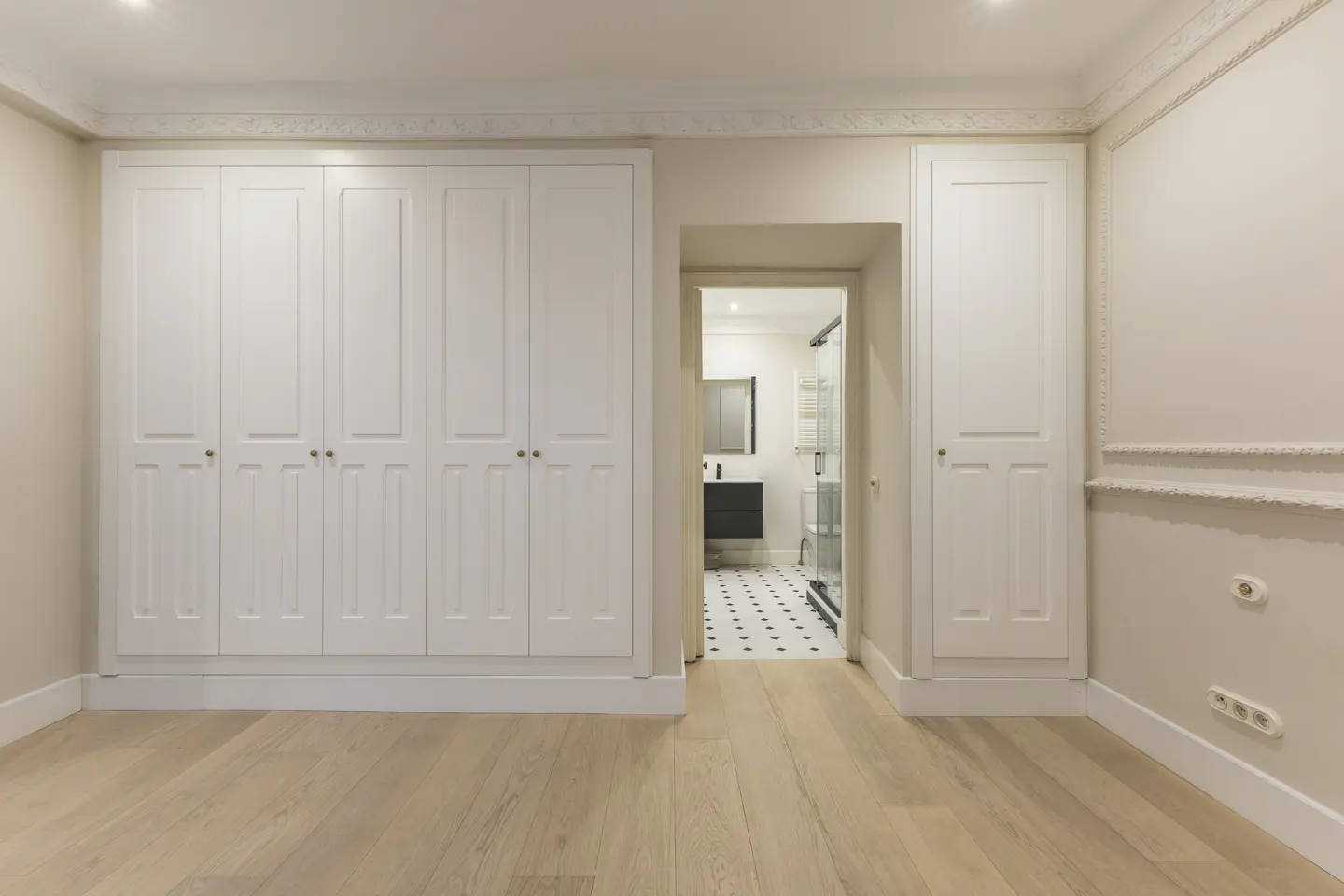 Bedroom with light wood floors, white walls, and built-in white wardrobes. A doorway leads to a bathroom with black and white tile.