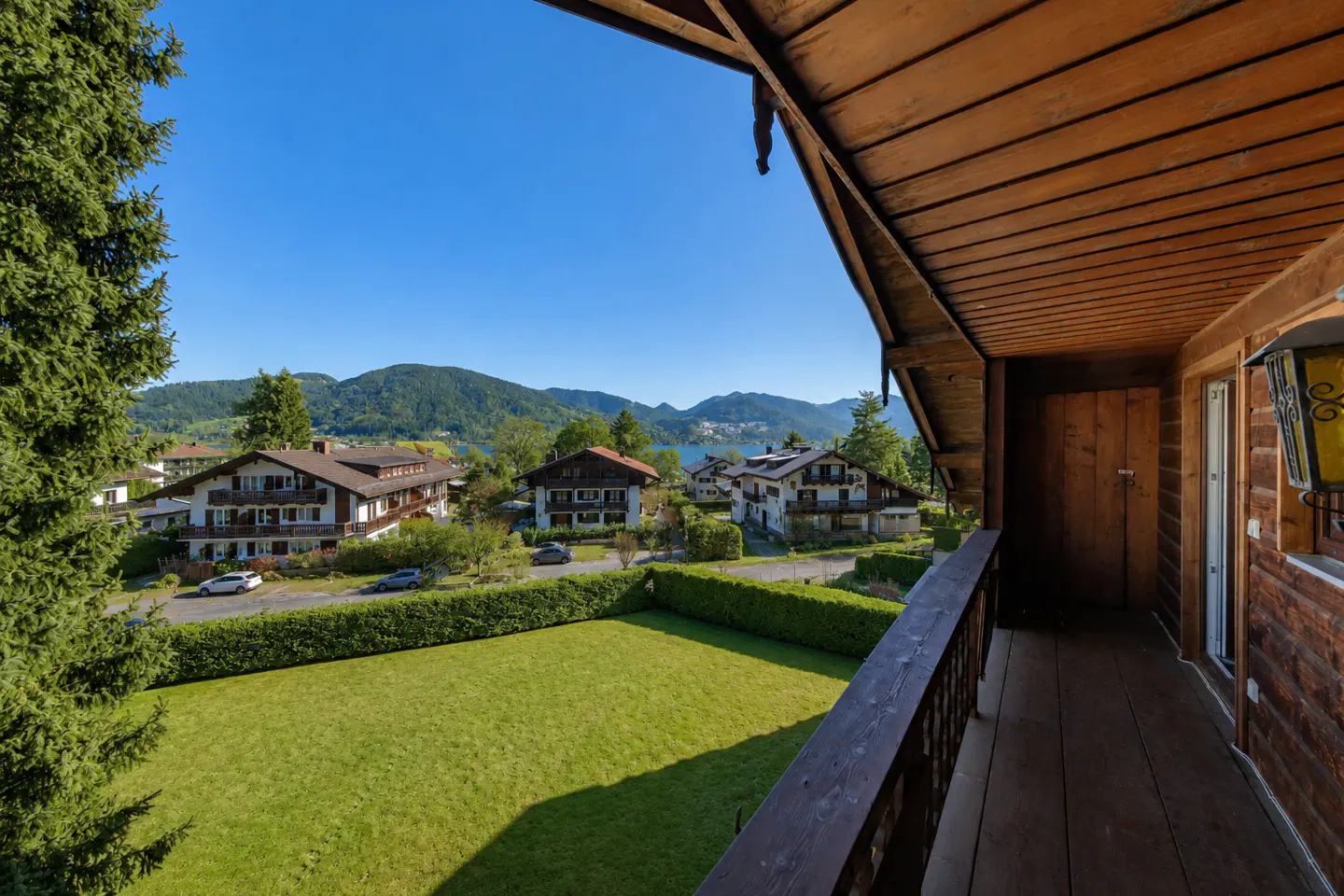 View from a wooden balcony overlooking a green lawn, traditional houses, and distant mountains under a clear blue sky.