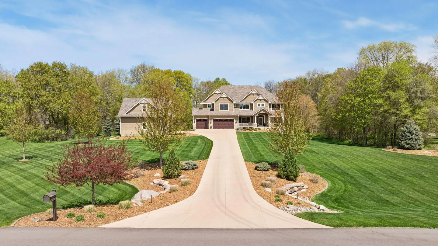 Two-story tan house with a long driveway and a well-manicured lawn on a sunny day.