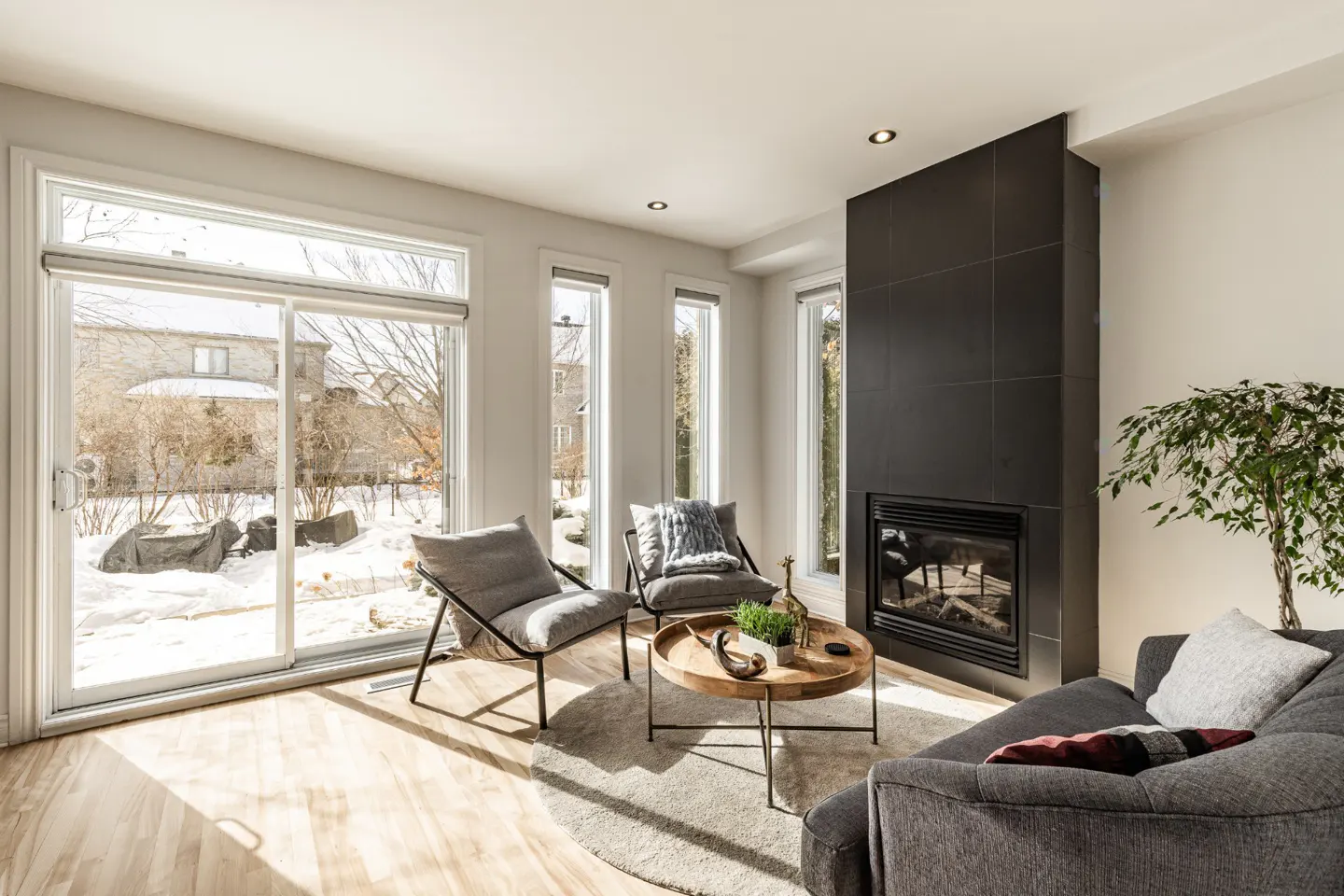Bright living room with wood floors, gray chairs, and a black fireplace. A sliding glass door shows a snowy yard.