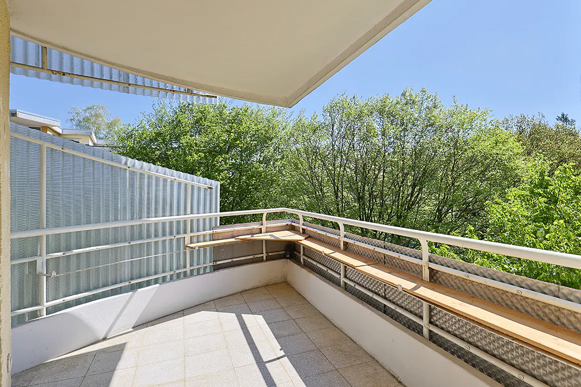 Balcony with white railings and tile floor overlooks green trees under a blue sky.