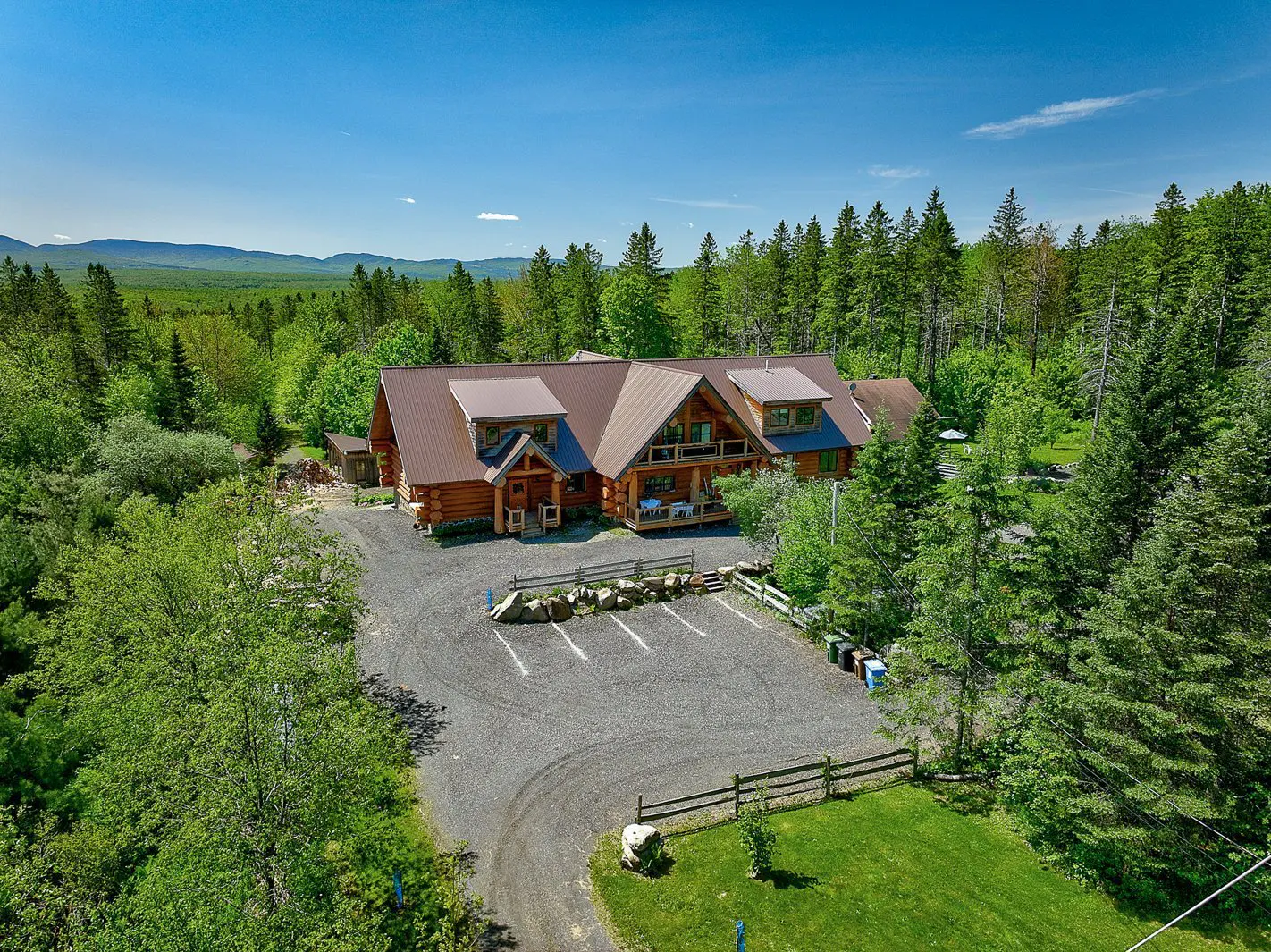 Aerial view of a large log cabin with a brown roof, surrounded by green trees and a gravel parking lot. Blue sky above.