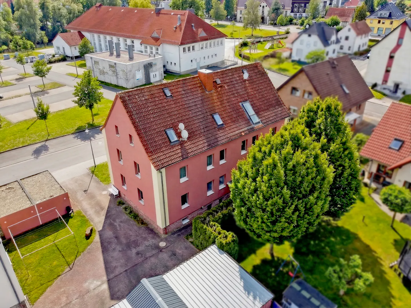 Aerial view of a three-story pink apartment building with a red tile roof, surrounded by green trees and other buildings.