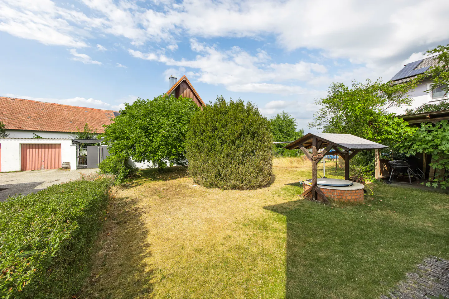 A backyard with a well, green trees, and a red-roofed building under a blue sky with white clouds.