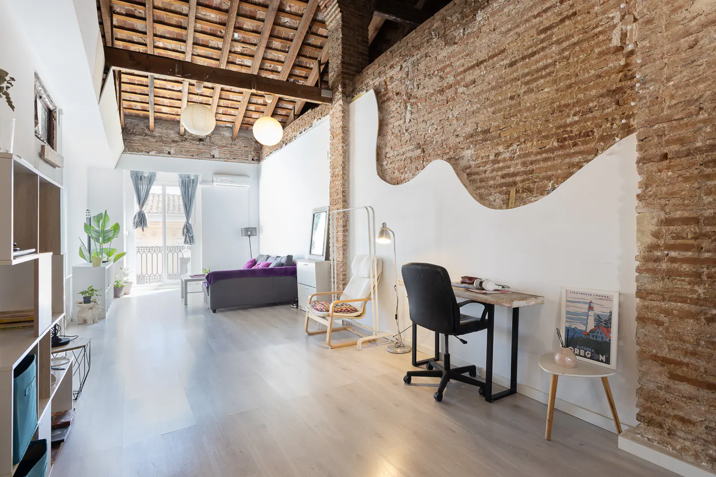 Bright, modern apartment interior with exposed brick wall, wooden ceiling beams, and light wood floors. A desk and sofa furnish the space.