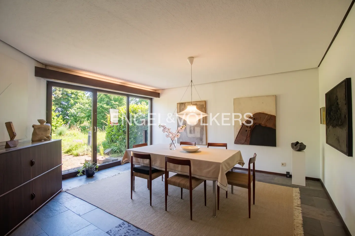 Dining room with a table set for six, beige tablecloth, and modern art on the walls. Sliding glass doors open to a green garden.