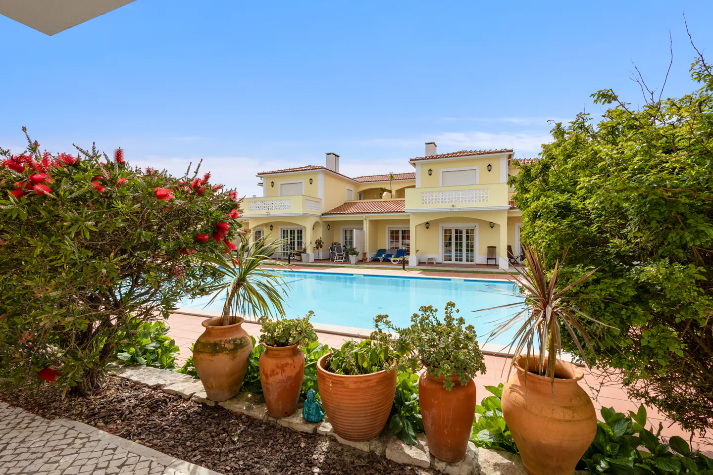 Exterior view of a yellow villa with a pool, terracotta pots, and lush greenery under a blue sky.