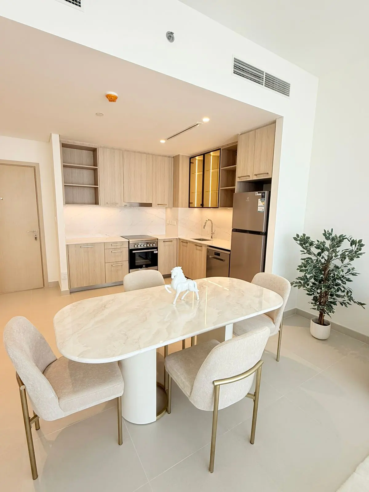 Modern kitchen with light wood cabinets, stainless steel appliances, and a marble dining table with four beige chairs.