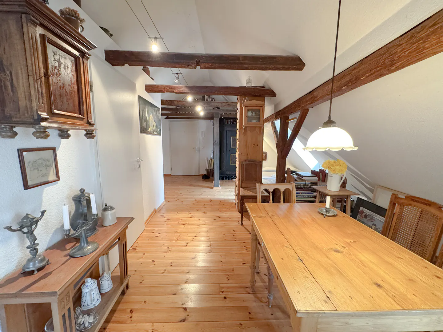 Attic room with wooden floors, exposed beams, and a long wooden table with chairs. A hallway leads to a door in the background.