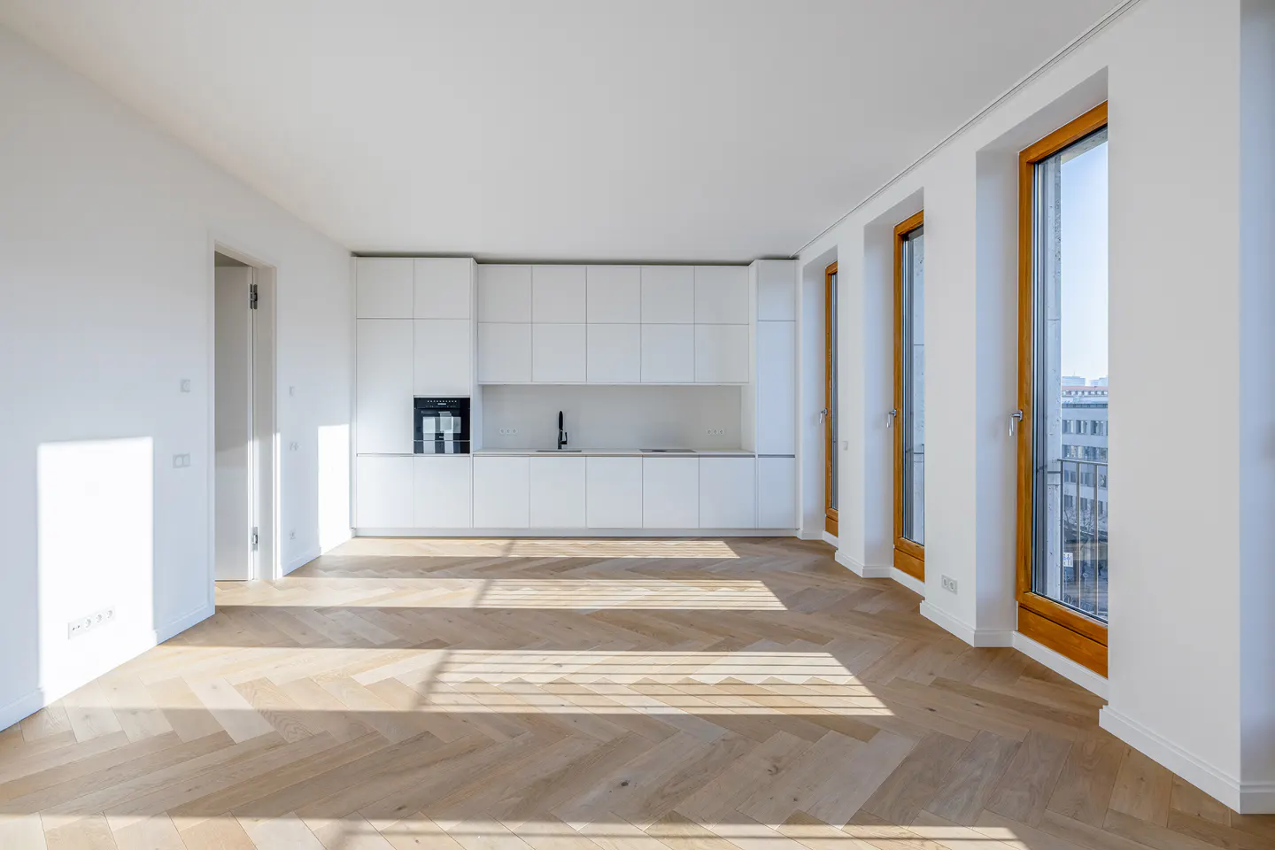 Bright, empty apartment with white walls, herringbone wood floors, and a modern white kitchen. Three large windows with wood frames let in sunlight.