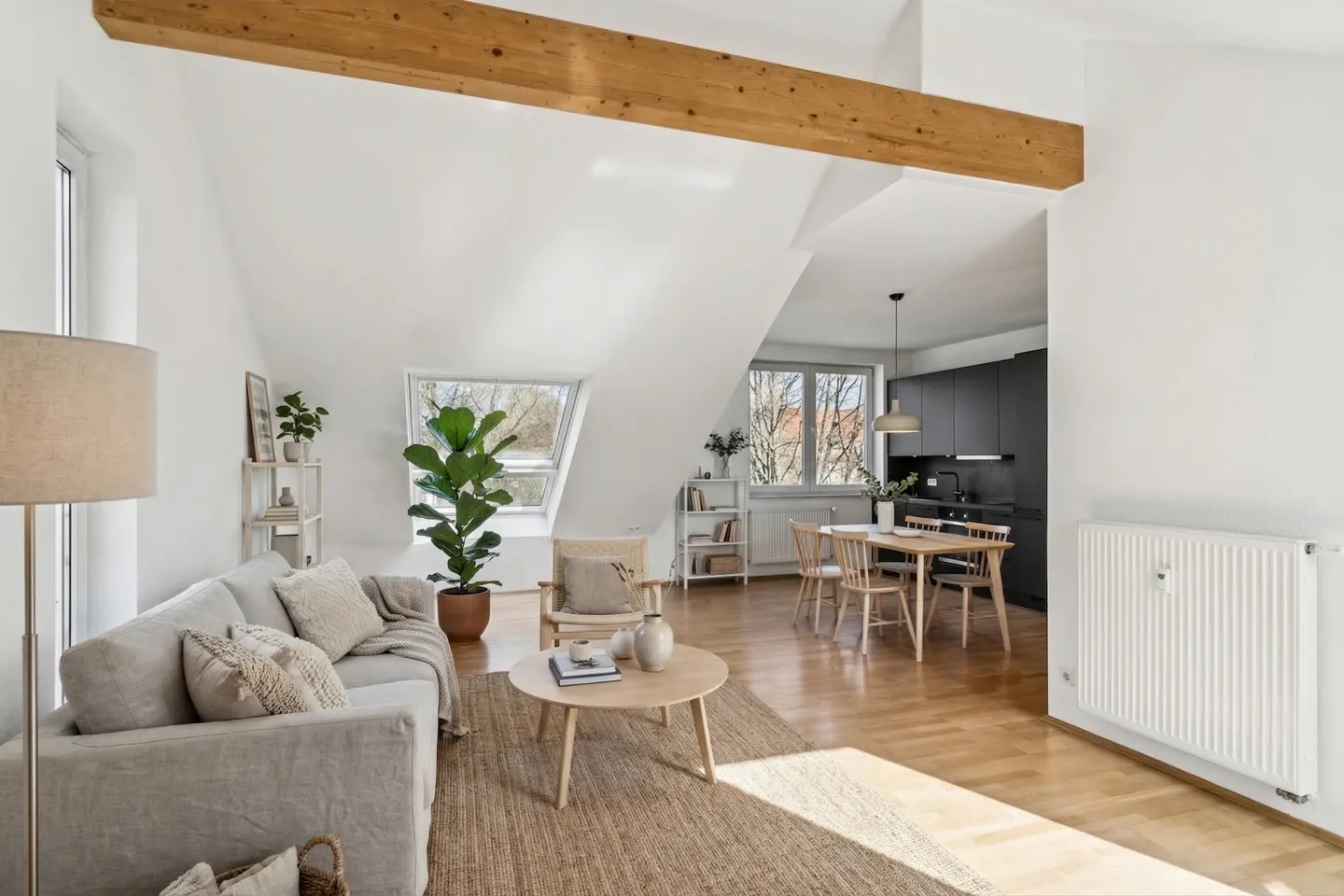 Bright, open-concept apartment with white walls, wood floors, and exposed beam. Living room with sofa, rug, and kitchen with dining table.