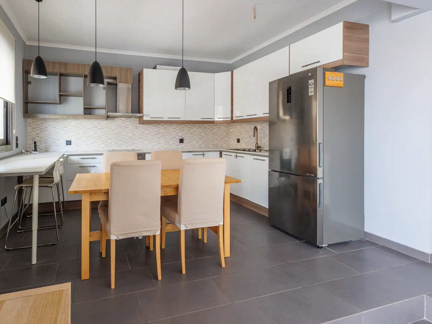 A modern kitchen with white cabinets, stainless steel refrigerator, and a wooden dining table with four chairs. Three black pendant lights hang above the table.