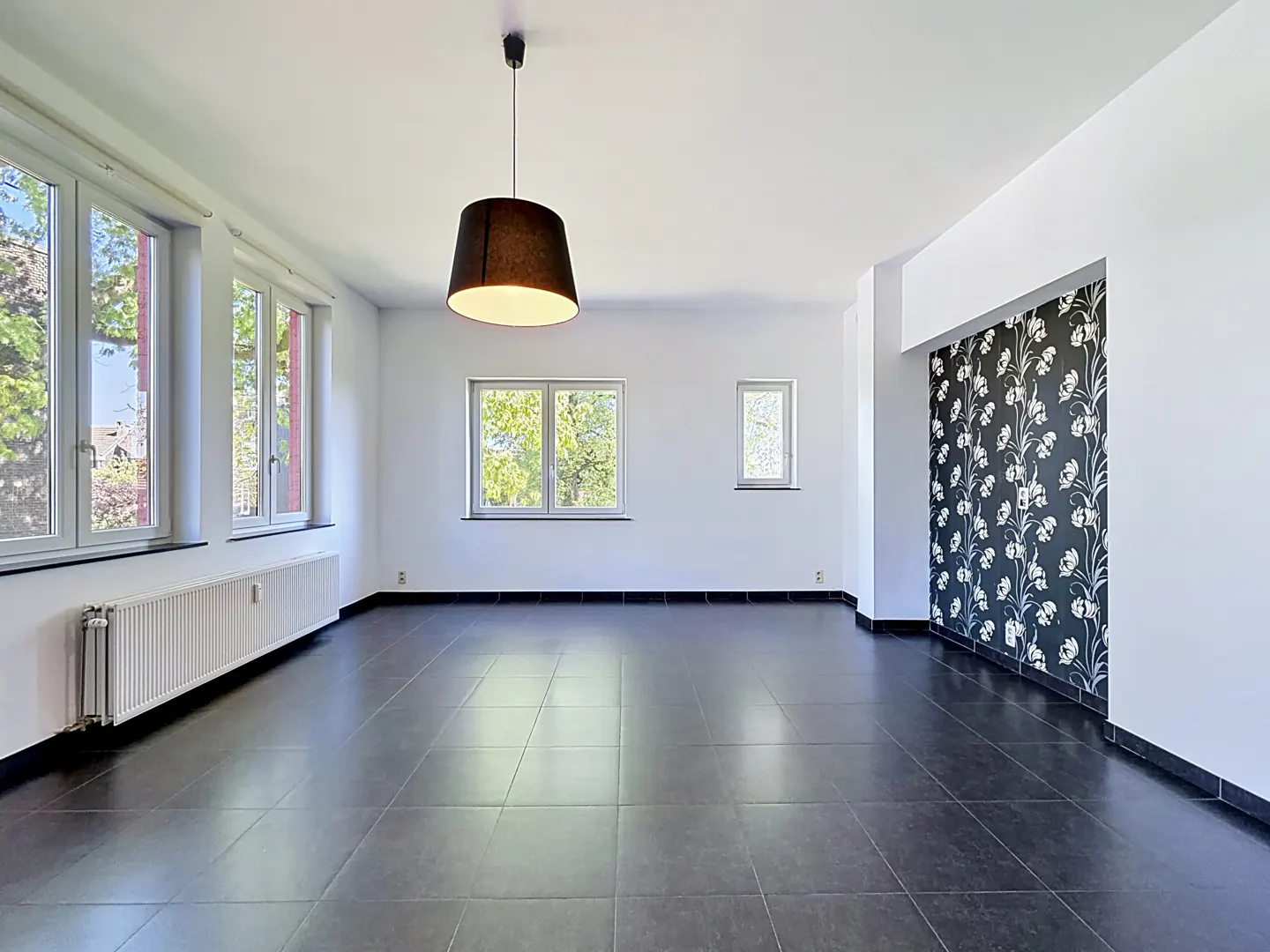 Bright, empty room with black tile floor, white walls, and large windows. A black lampshade hangs from the ceiling. Floral wallpaper accents one wall.