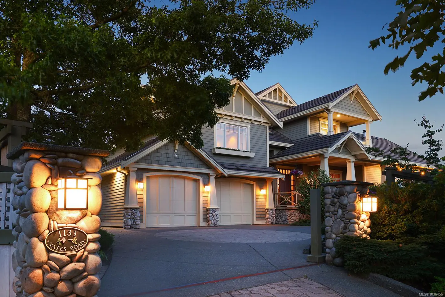 Two-story house with gray siding, white trim, and a two-car garage at dusk. Stone pillars flank the driveway. Address plaque reads "1133 Yates Road."
