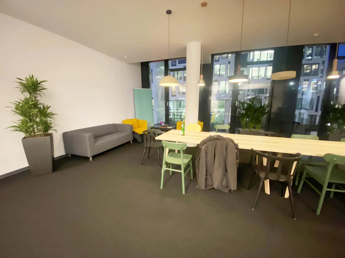 Office space with a long table, chairs, gray sofa, yellow chair, and potted plant. Large windows show city buildings.