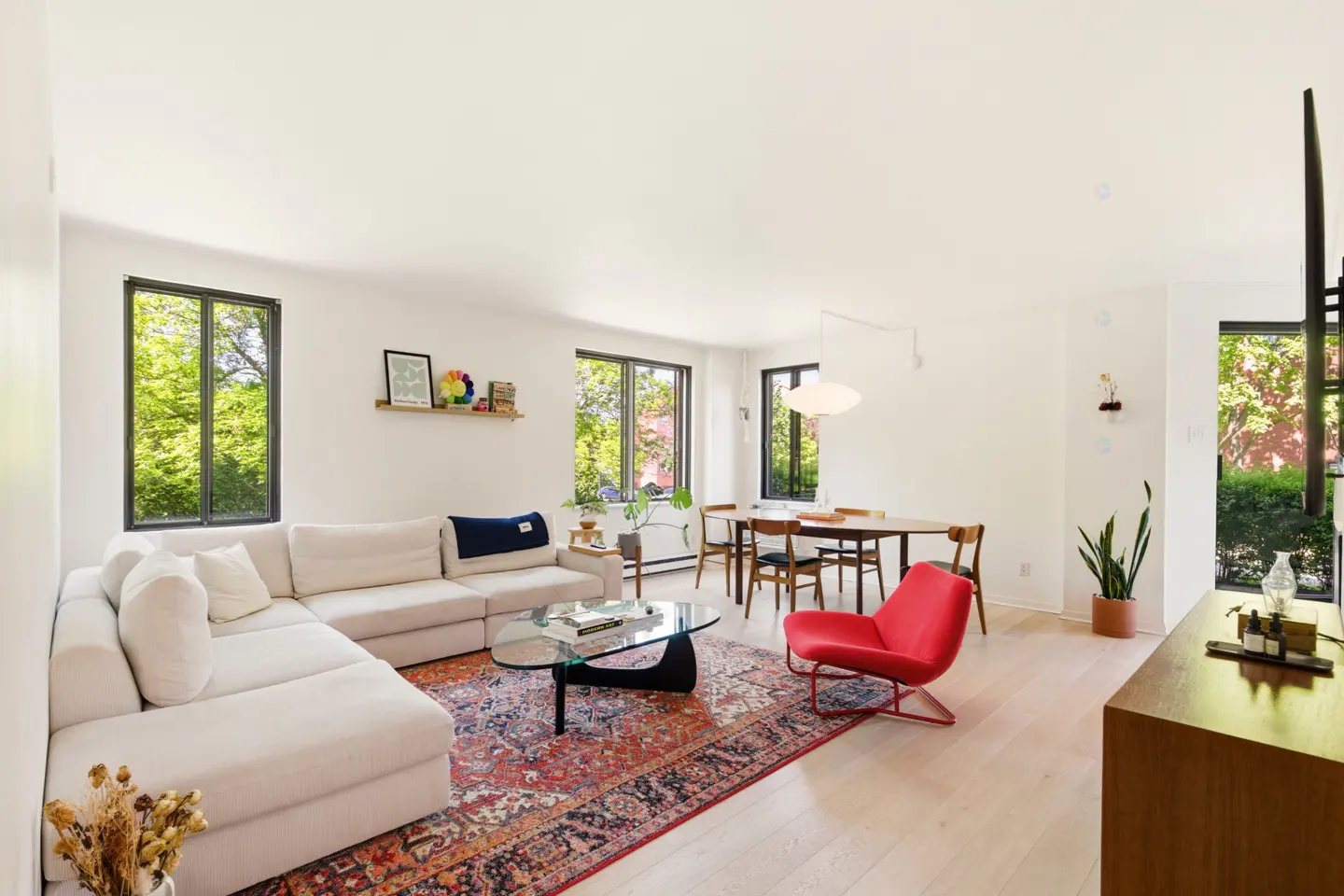 Bright living room with white walls, light wood floors, and black-framed windows. A white sectional sofa faces a glass coffee table on a patterned rug. A red chair sits near a wood dining table.