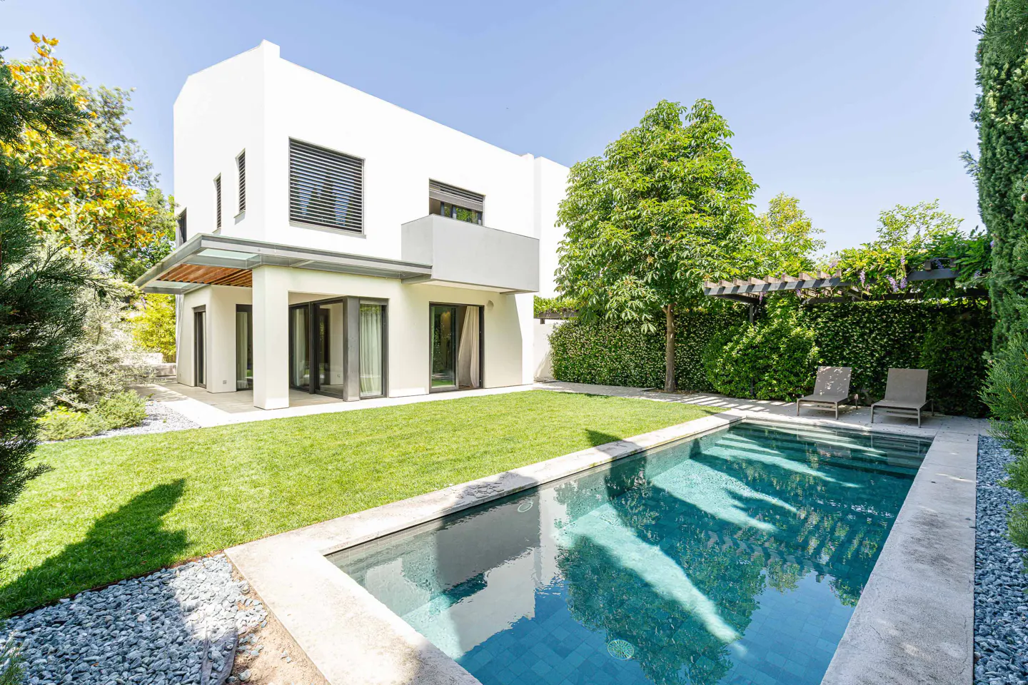 Modern white house with a pool. The backyard has green grass, trees, and two lounge chairs.