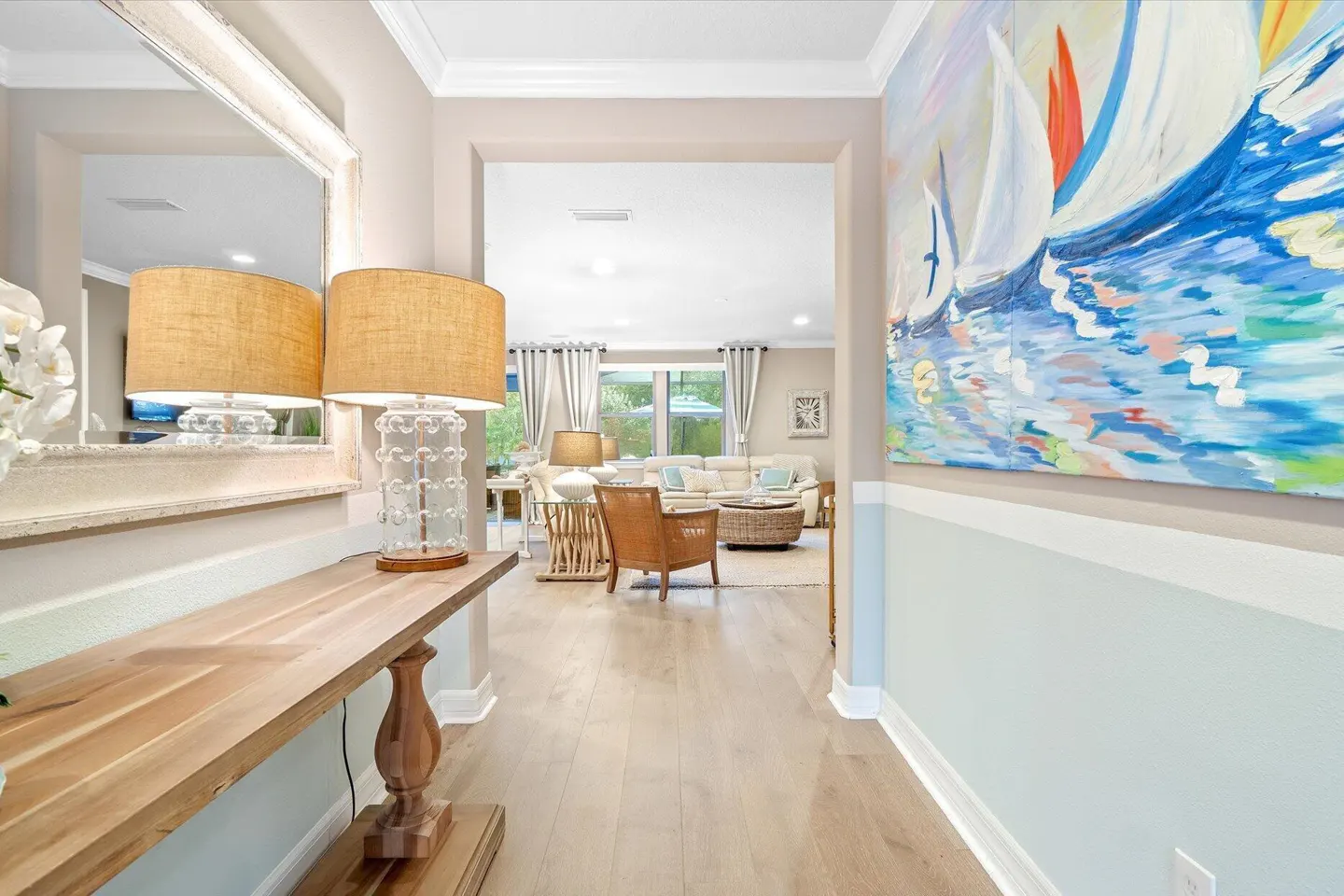 Hallway with a wooden console table, lamp, and large sailboat painting, leading to a bright living room with seating.