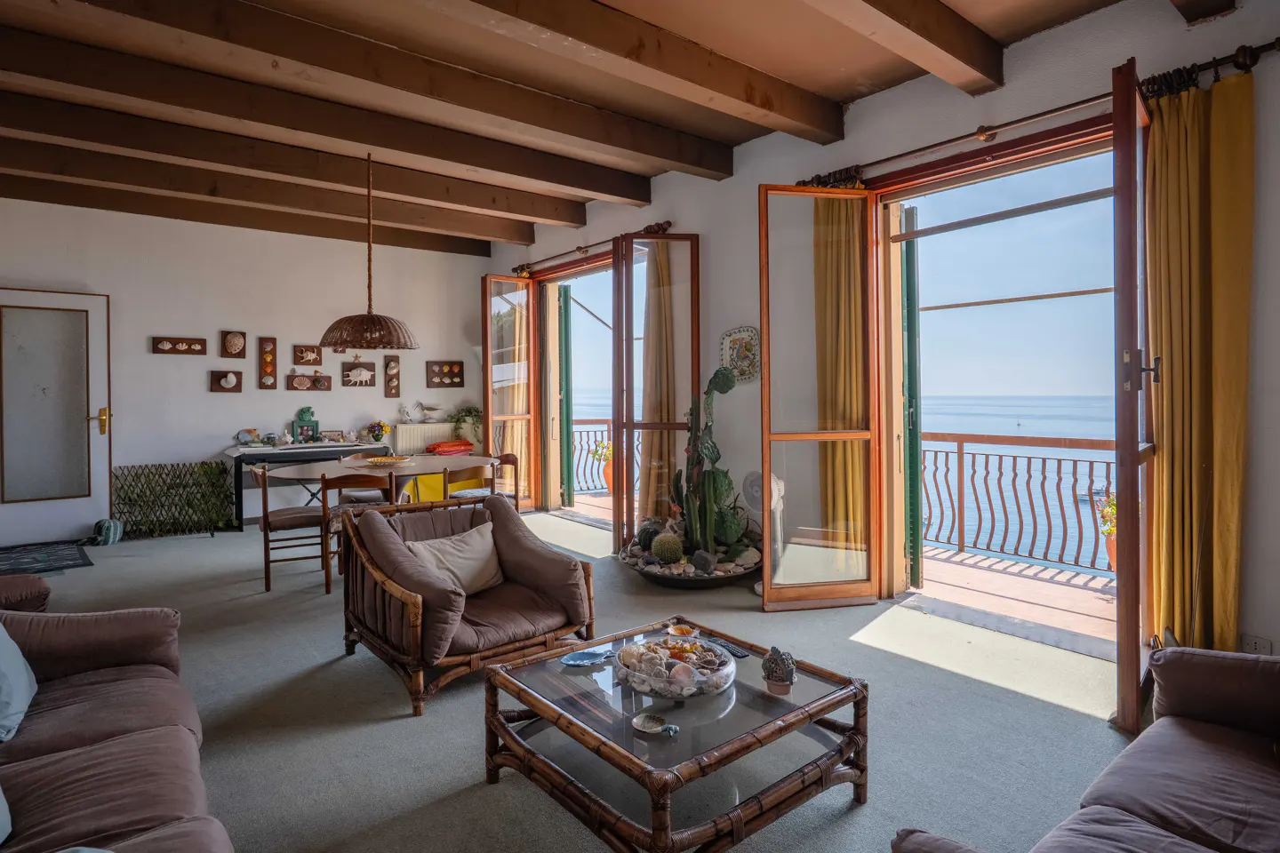 Living room with brown furniture, exposed wood ceiling beams, and open doors leading to a balcony overlooking the ocean.