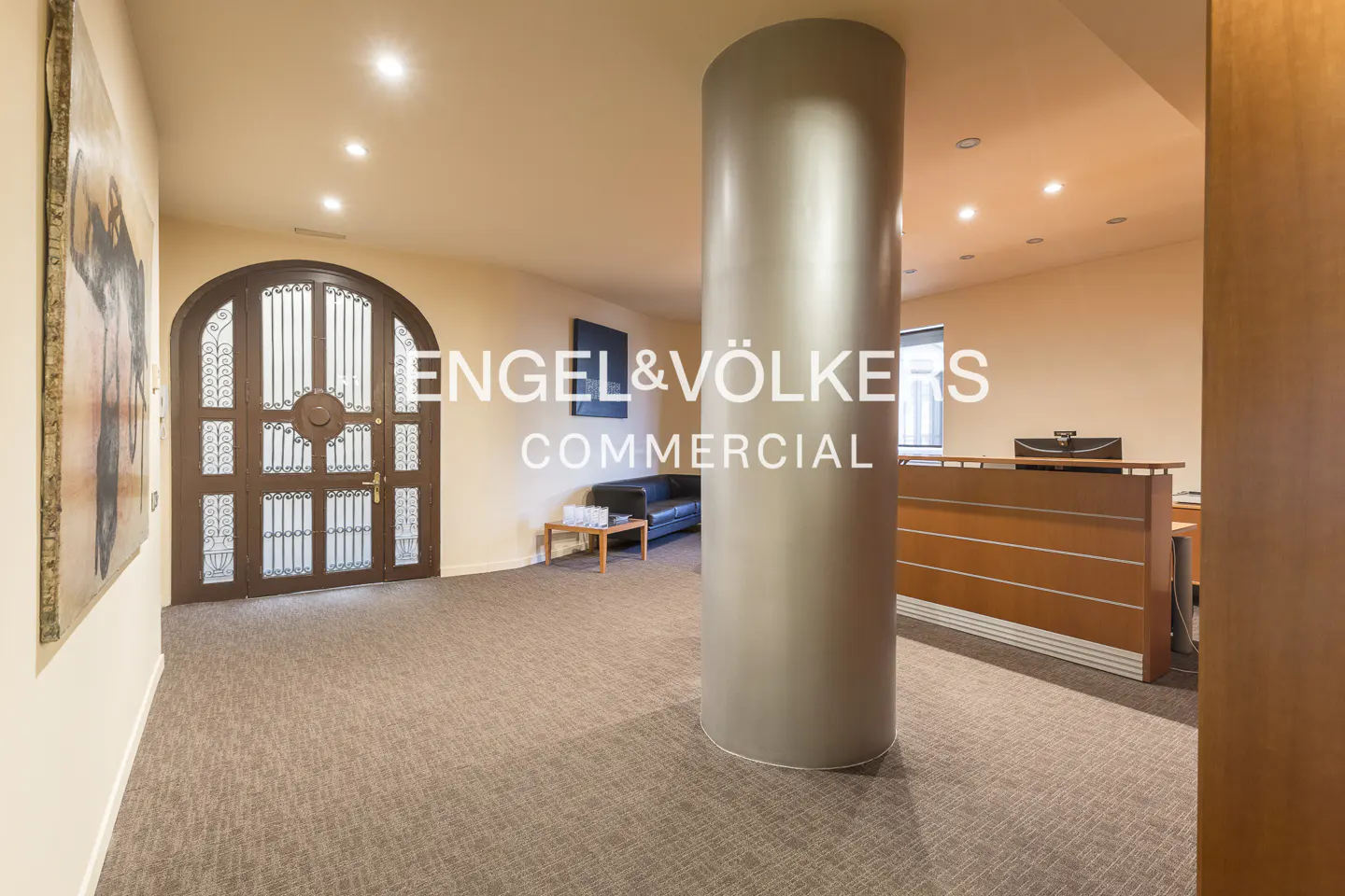 Office lobby with a large gray column, reception desk, and arched wooden door. A black sofa sits in the background.