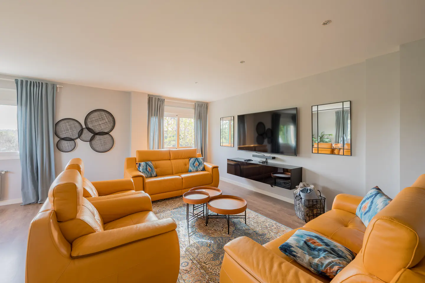 Bright living room with tan leather sofas, blue patterned pillows, and a patterned rug. A large TV hangs above a black console.
