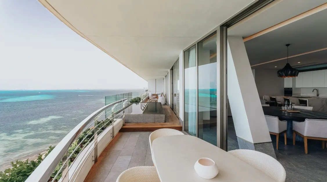 Balcony view of ocean. White table and chairs on stone patio. Glass railing. Open sliding glass doors to dining room and kitchen.