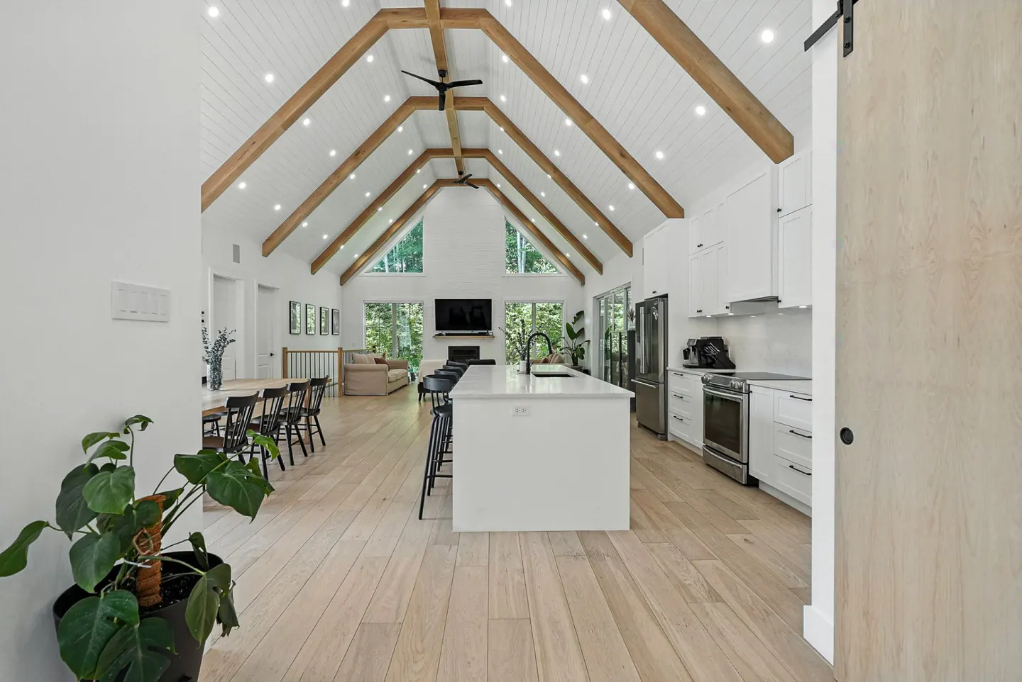 Bright, open-concept kitchen and living space with vaulted ceiling, wood beams, white cabinets, and a large island with black stools.