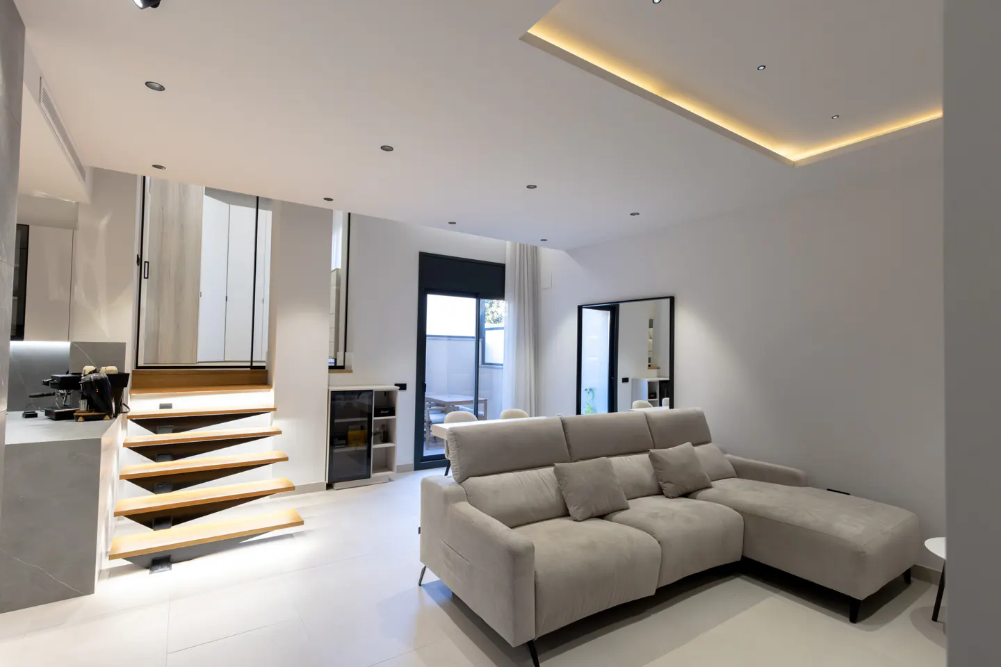 Bright, modern living room with a beige sectional sofa, white walls, and floating wooden stairs leading up. A black-framed door opens to a patio.