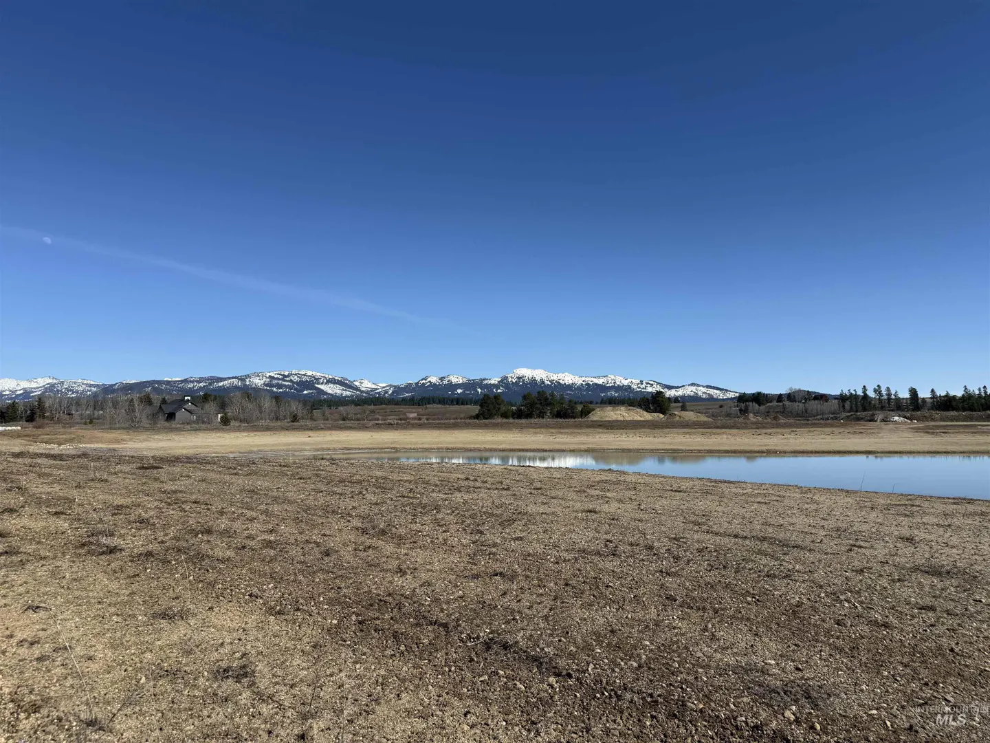 Scenic view of a brown field with a small pond, snow-capped mountains, and a clear blue sky.