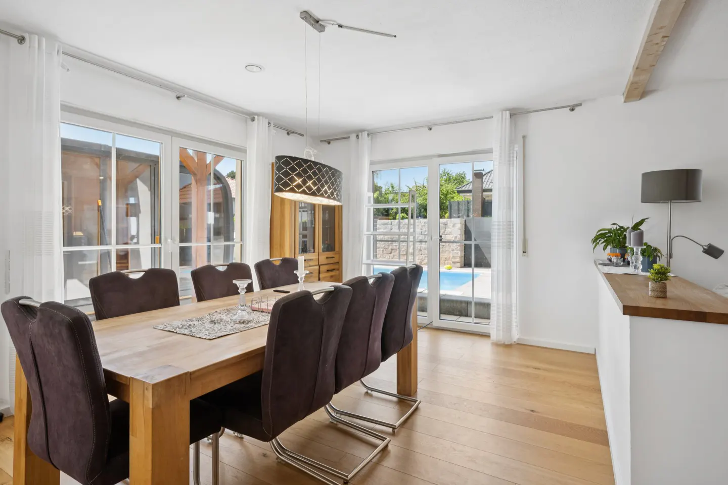 Dining room with a wooden table, dark chairs, and a modern pendant light, overlooking a patio with a swimming pool.
