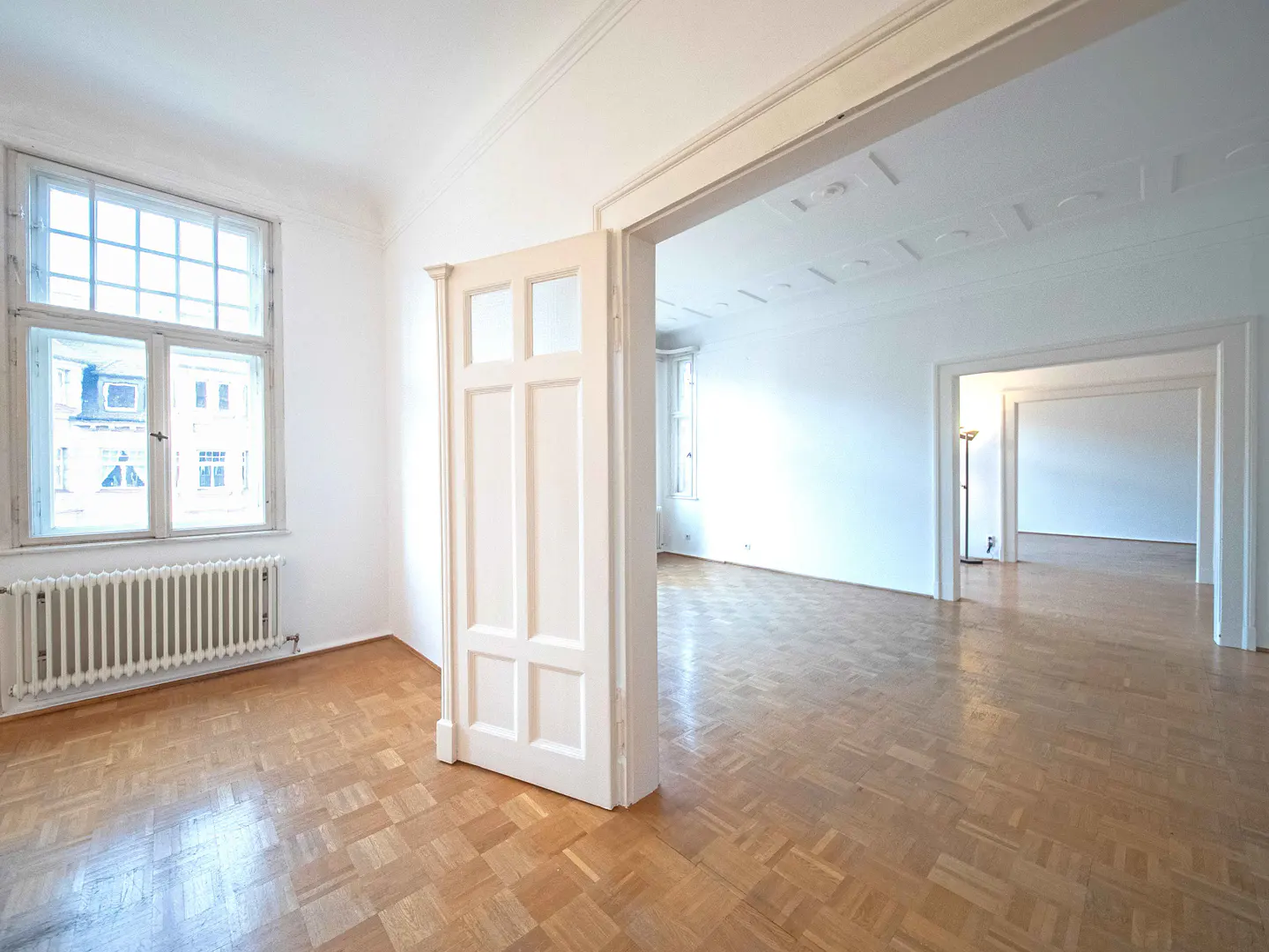 Bright, empty room with parquet floors, white walls, and an open white door leading to other rooms. A window with a radiator is on the left.