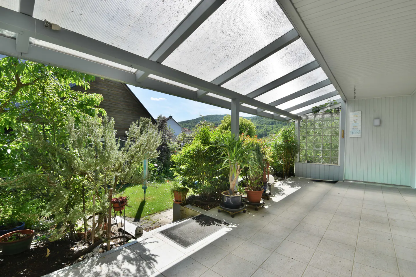 Covered patio with light-colored tile flooring, potted plants, and a view of a green garden and distant hills.