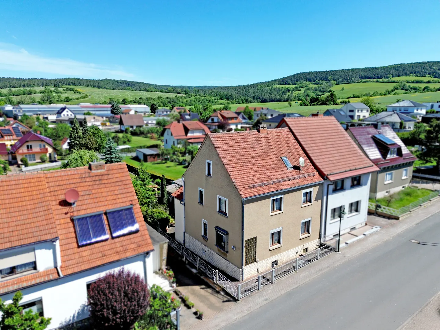 Aerial view of a tan house with a red tile roof in a quiet residential neighborhood with green hills in the background.