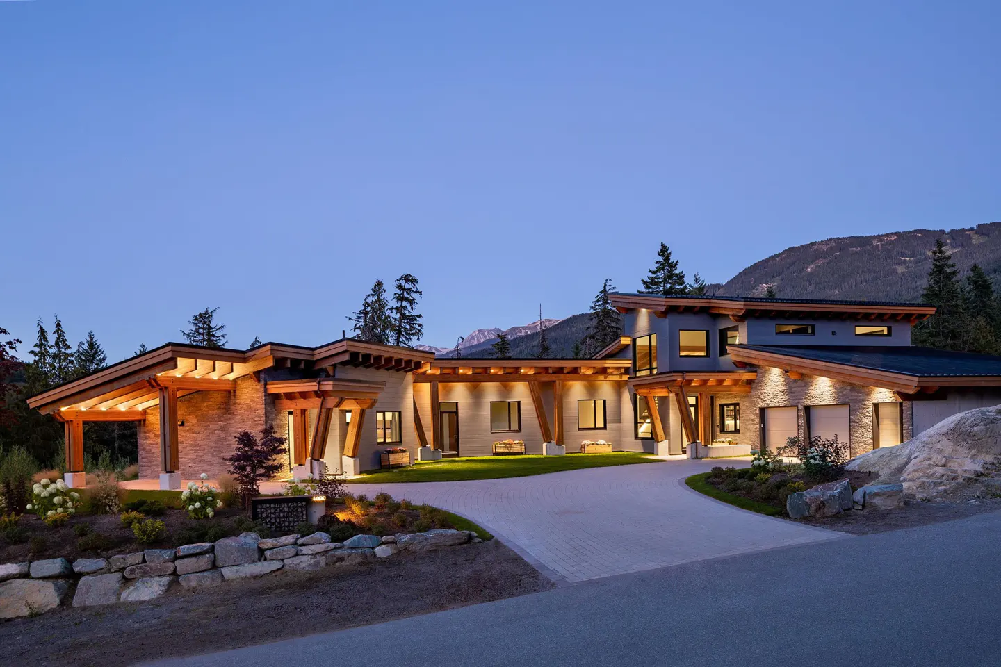 A modern, single-story home with wood beams and stone accents, set against a mountain backdrop at dusk. A paved driveway leads to the house.