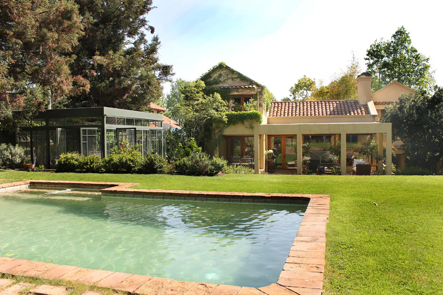 A backyard with a pool, a greenhouse, and a house with a red tile roof.