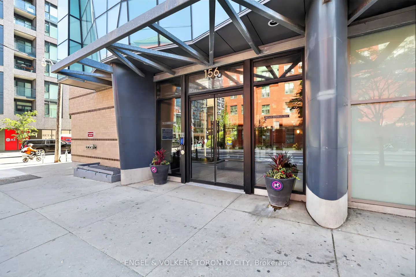 Modern glass entrance of a Toronto condo building at 156, featuring dark metal framing and two large potted plants.