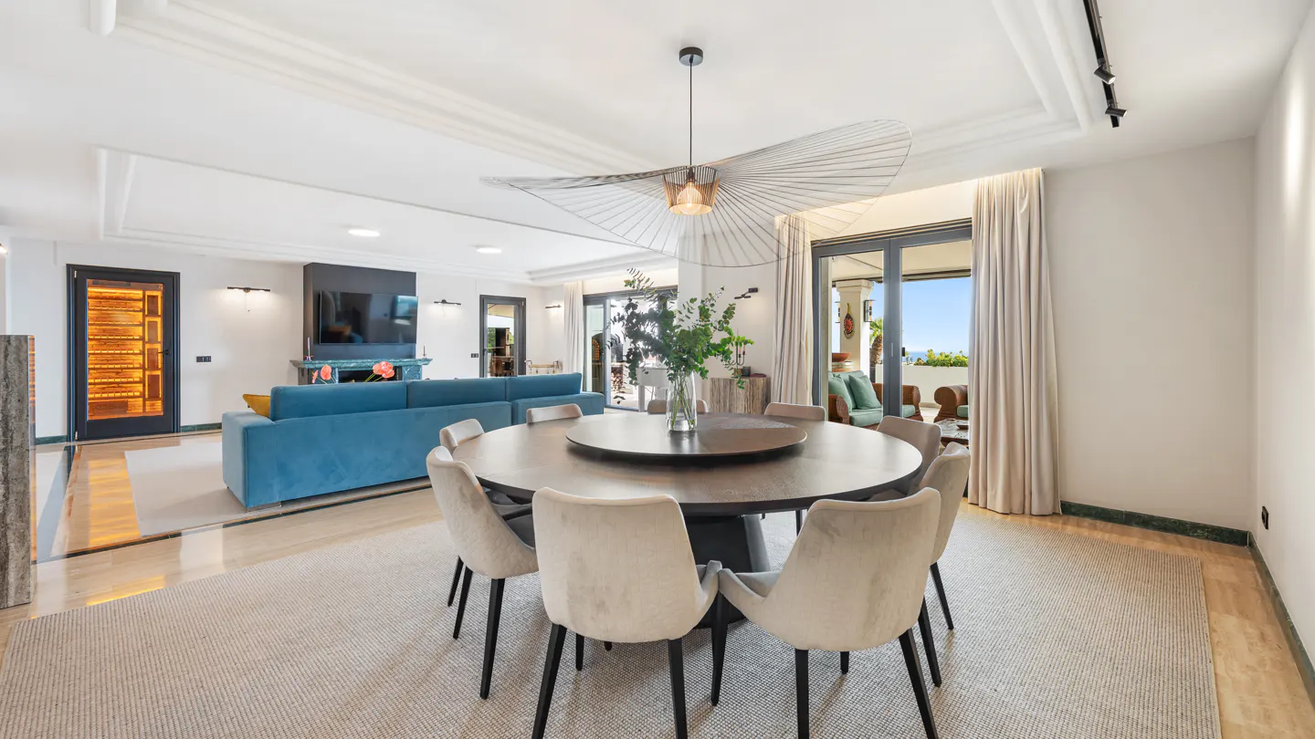 Bright, modern living and dining area with a round table, beige chairs, and a blue sofa. A unique light fixture hangs above the table. Balcony view in the background.