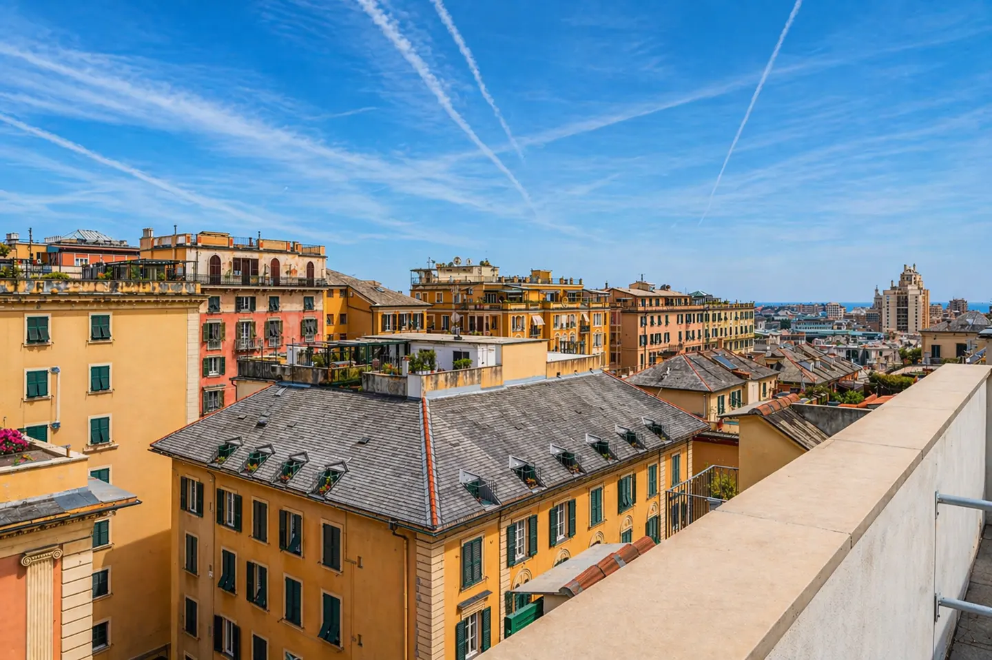 View of Genoa, Italy, from a rooftop. Buildings are yellow, orange, and pink with green shutters. Blue sky with contrails.