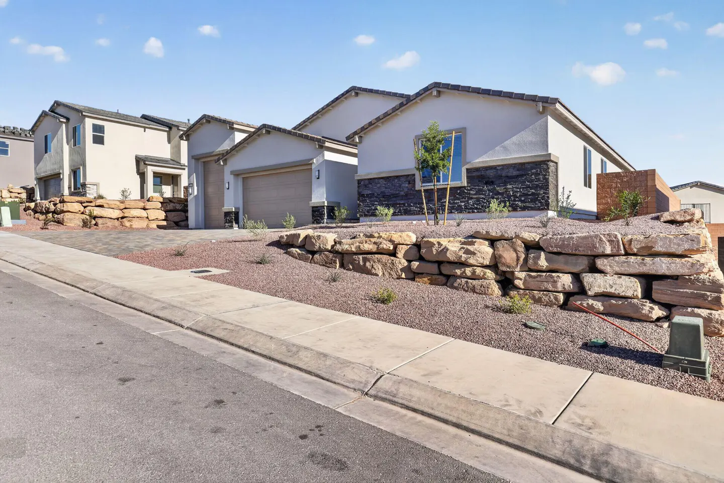 A modern, single-family home with a beige exterior and a stone accent wall, set on a landscaped lot with a rock retaining wall.