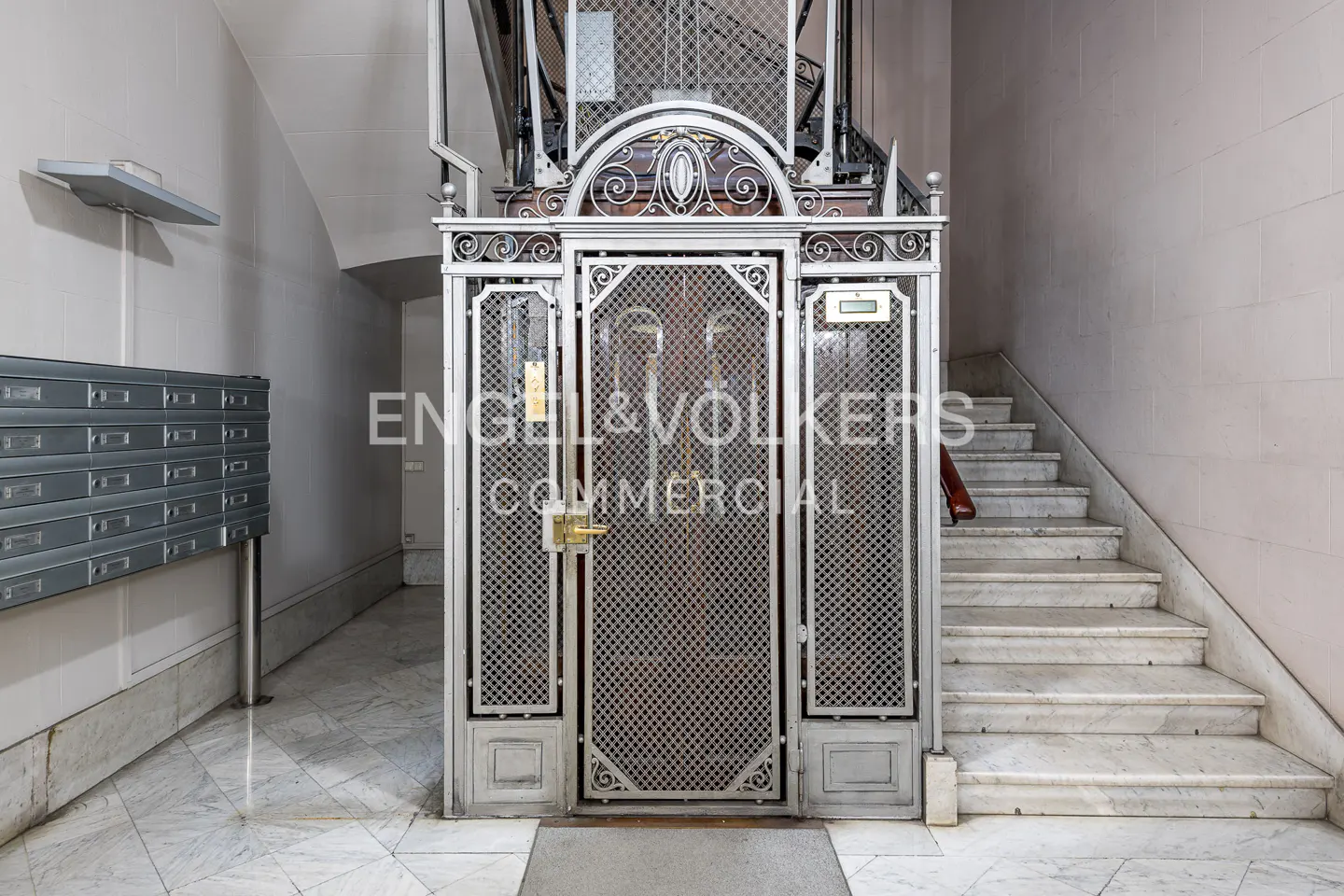 Interior view of a vintage metal elevator with a lattice door, next to marble stairs and mailboxes in a building lobby.