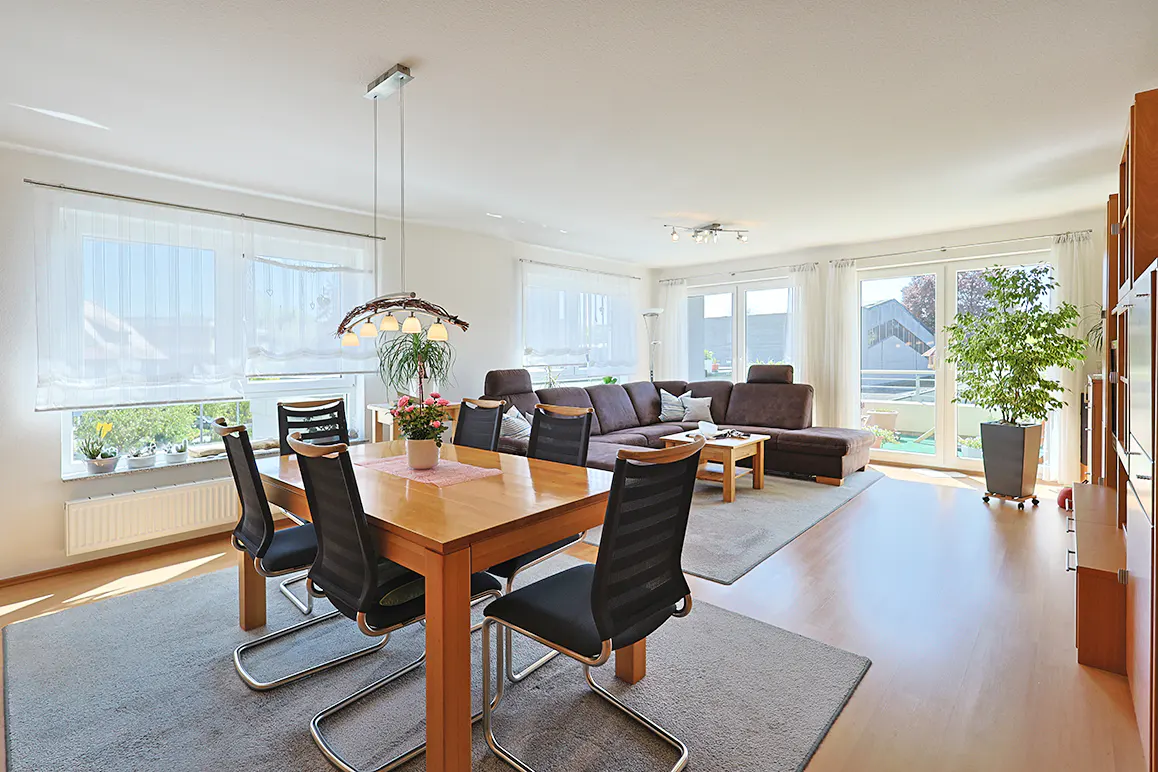 Bright open-plan living and dining room with a wooden table, black chairs, brown sectional sofa, and large windows.