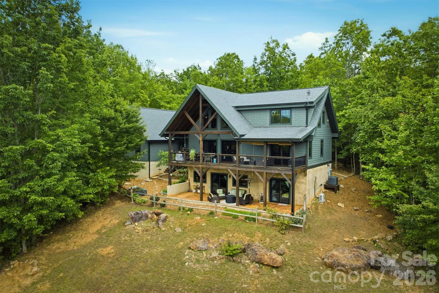 Two-story gray house with wooden beams and balconies, surrounded by green trees and a grassy lawn.