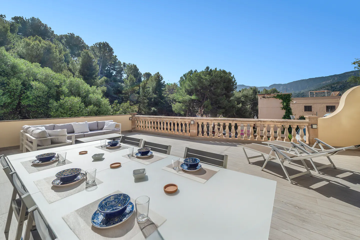 Outdoor terrace with a long white table set for dining, a sofa, and lounge chairs. Balustrade overlooks lush green trees and a clear blue sky.