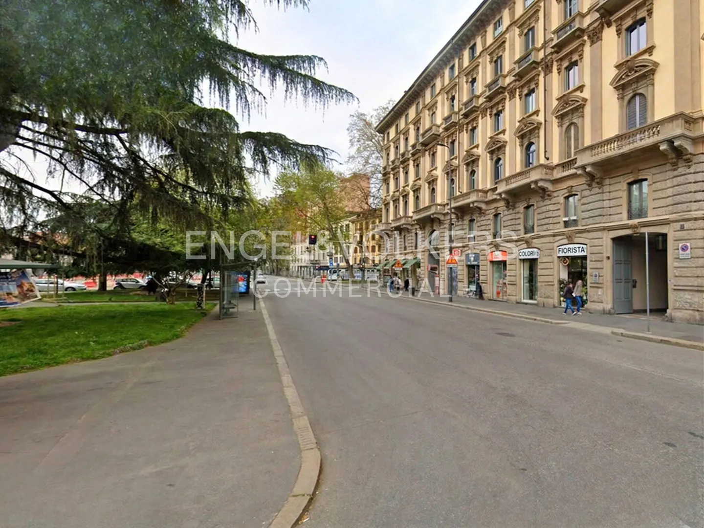 Street view of a European city with a park on the left and shops on the right. The building is tan with many windows.