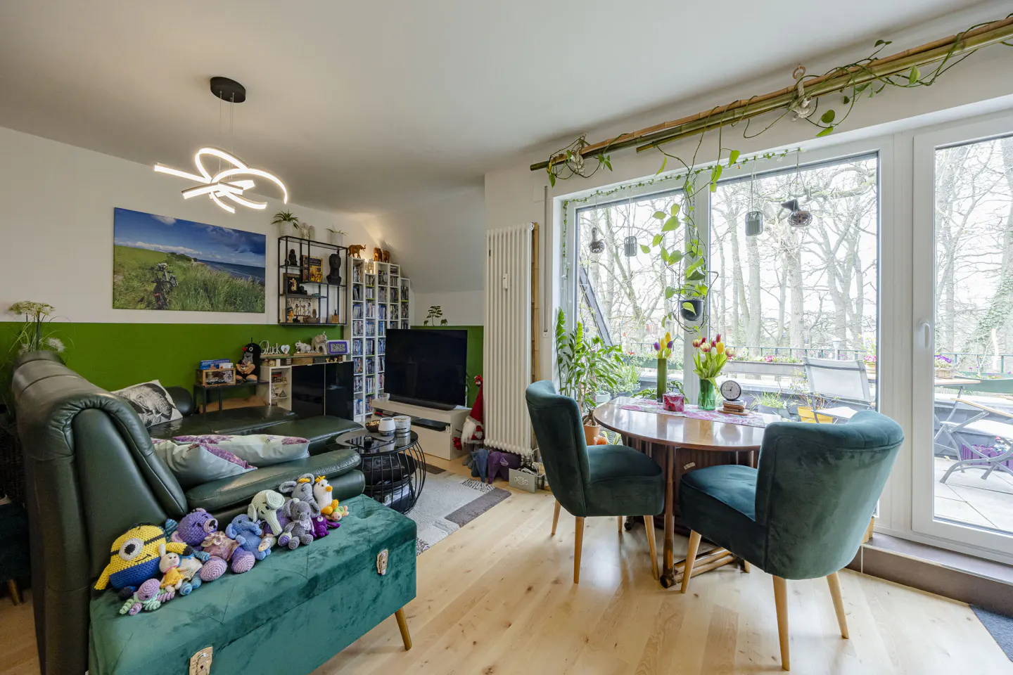 Bright living room with green sofa, ottoman with stuffed animals, and round table with chairs near a large window.